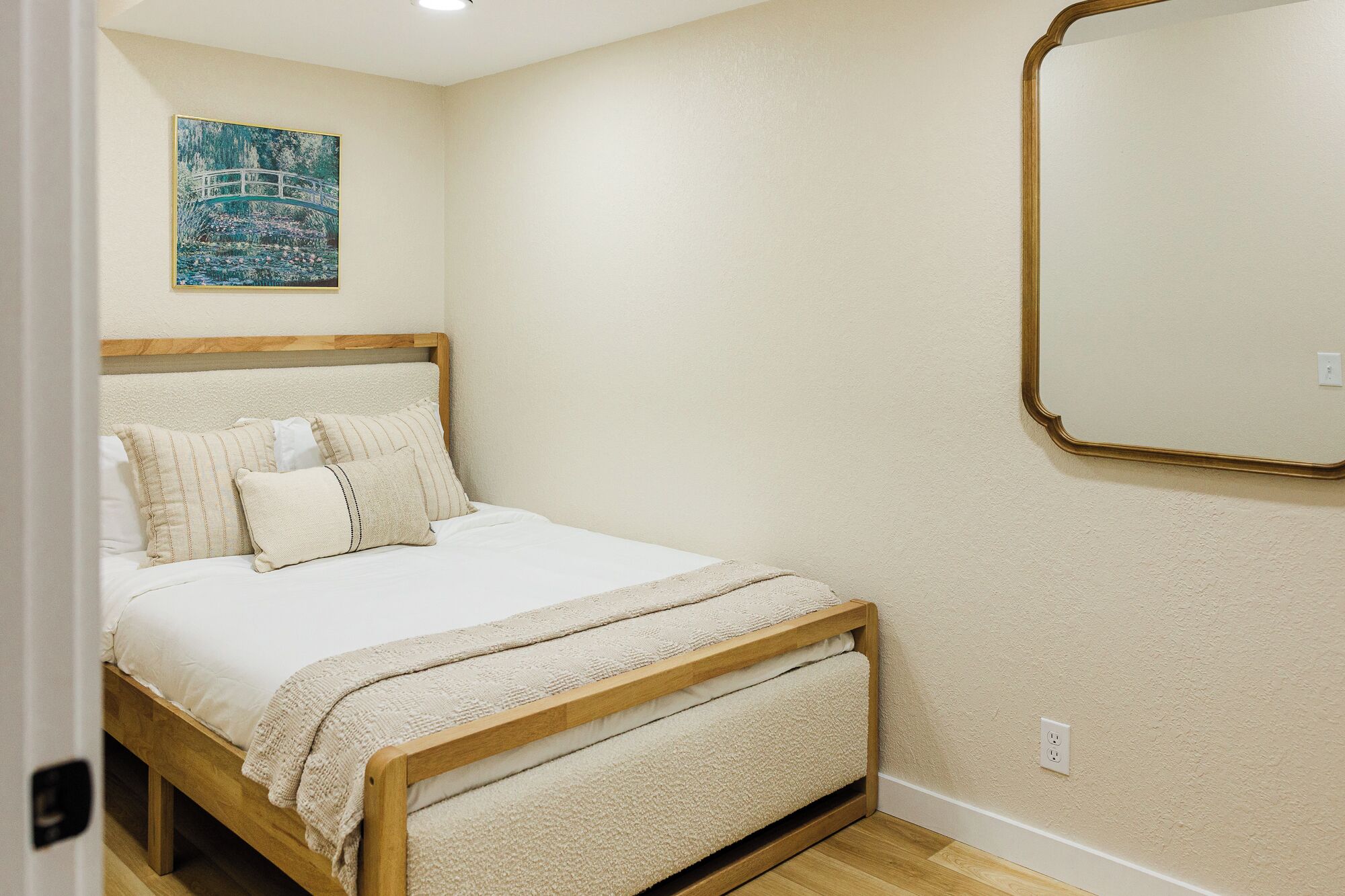 A bedroom with a bed, pillows, a blanket, a wooden headboard, a wall-mounted mirror, and a painting.