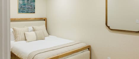 Un dormitorio con cama, almohadas, una colcha, cabecera de madera, un espejo en la pared y un cuadro.