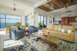 A spacious living area with a large window offering a view of the sea, a beige sofa, a blue armchair, and a glass coffee table.