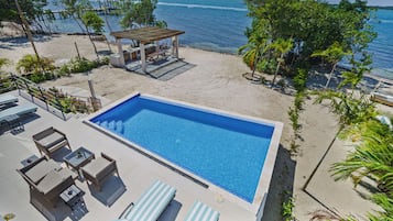 A rectangular swimming pool with lounge chairs and a covered seating area on a sandy beach, overlooking the sea.