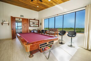A pool table with a red felt top, surrounded by two black bar stools, in a room with a wooden ceiling and large windows offering a view of greenery.