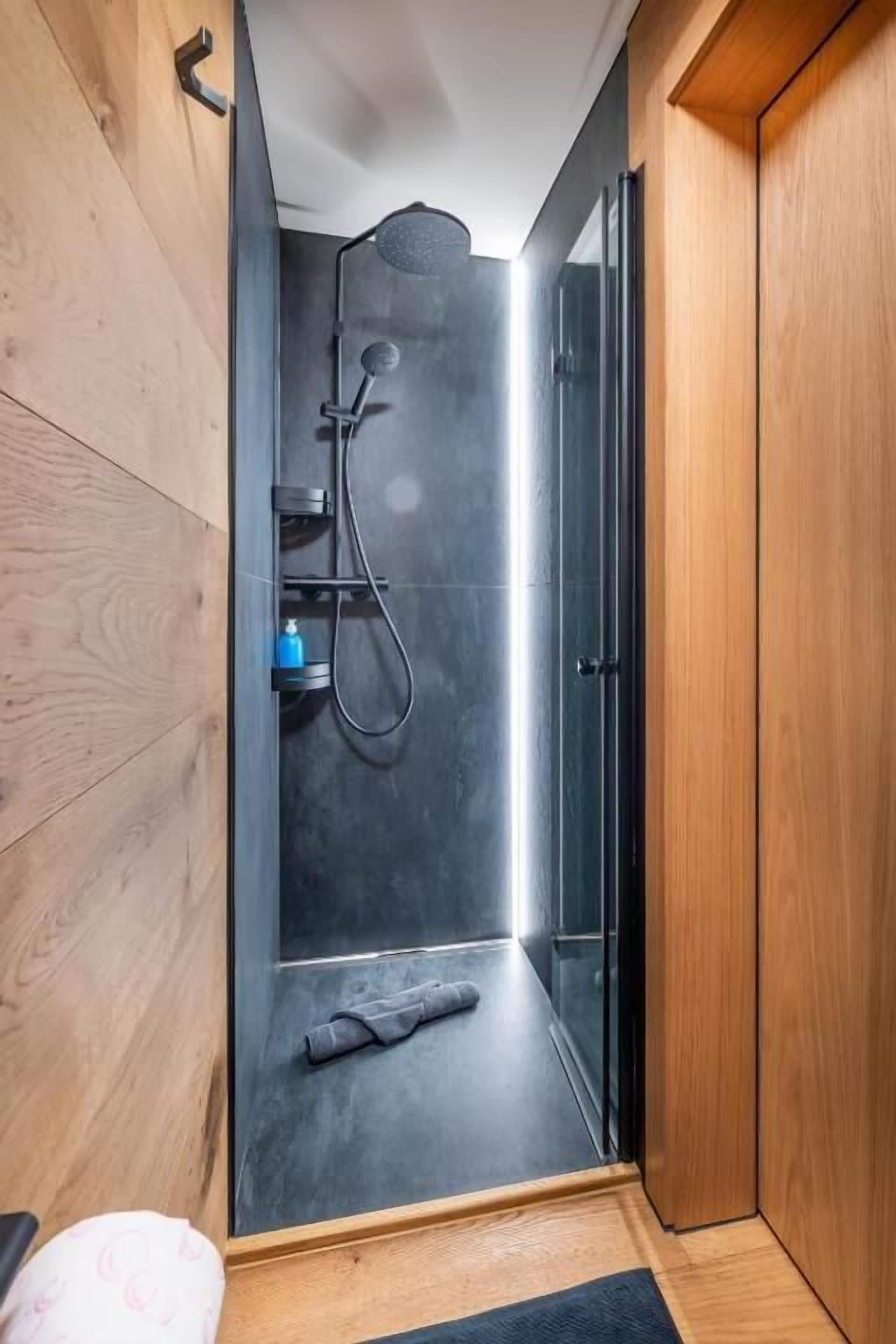 Una doccia con porta in vetro, un asciugamano sul pavimento e una parete in legno.