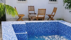 Piscina rectangular con mosaicos azules, rodeada por un muro blanco con ventanas y sillas de madera.