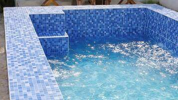 En rektangulär swimmingpool med blå mosaikplattor, omgiven av en vit vägg med fönster och trämöbler.