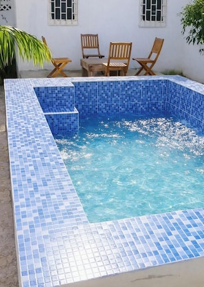Una piscina rettangolare con piastrelle di mosaico blu, circondata da un muro bianco con finestre e sedie di legno.