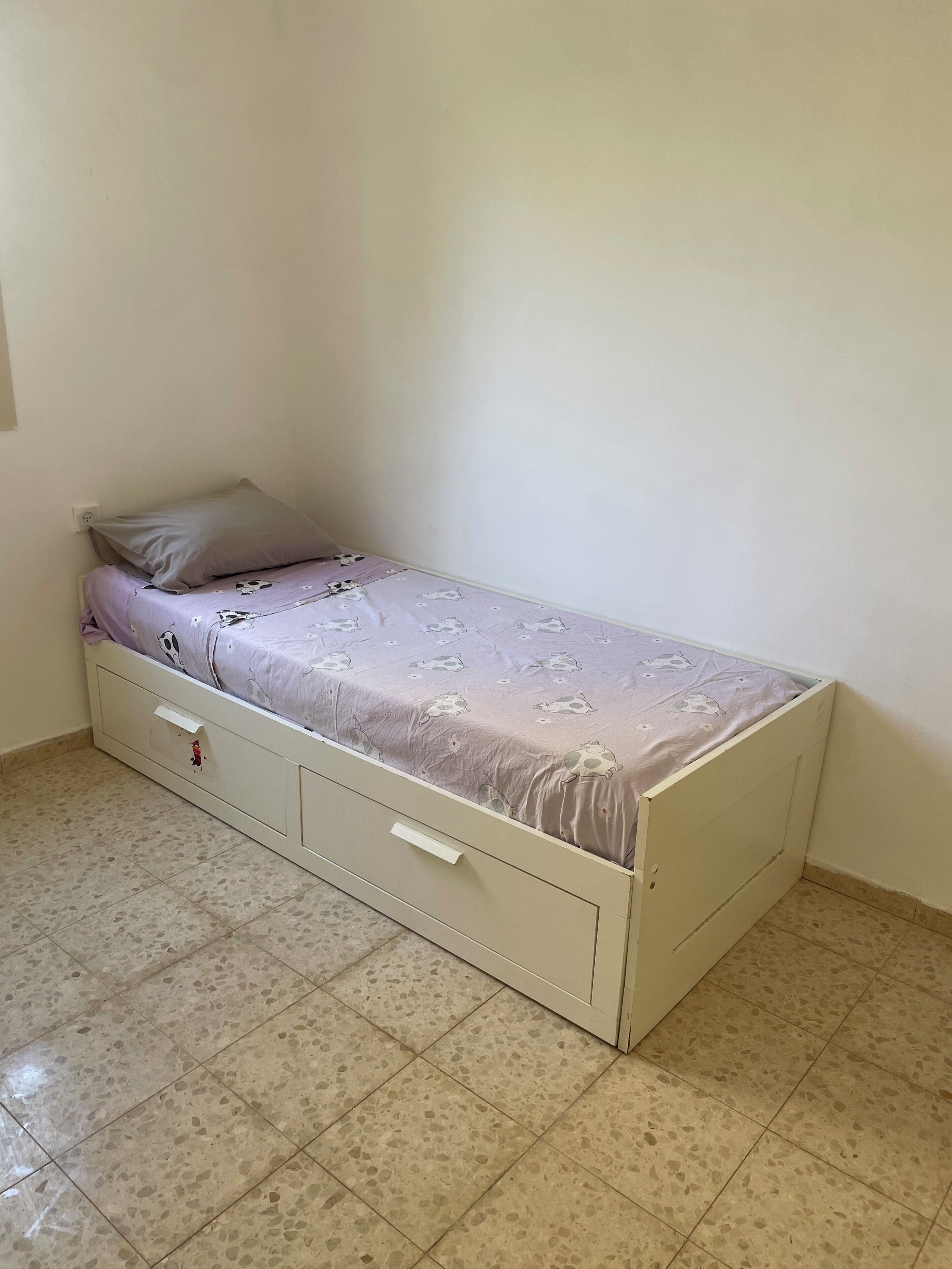 Une petite chambre avec un lit simple et un aménagement sobre, comprenant un lit recouvert d’une couvre-lit à motifs, une petite commode et un mur uni.