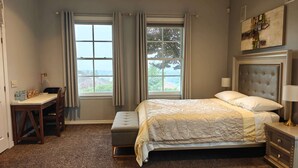 A bedroom with a bed, bedside table, lamp, and a desk with a chair. There are two windows with curtains, and a painting on the wall.