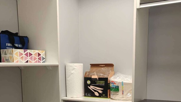 A kitchen with a black refrigerator, a white cabinet with a blue bag, and a shelf with a box and a roll of paper towels.