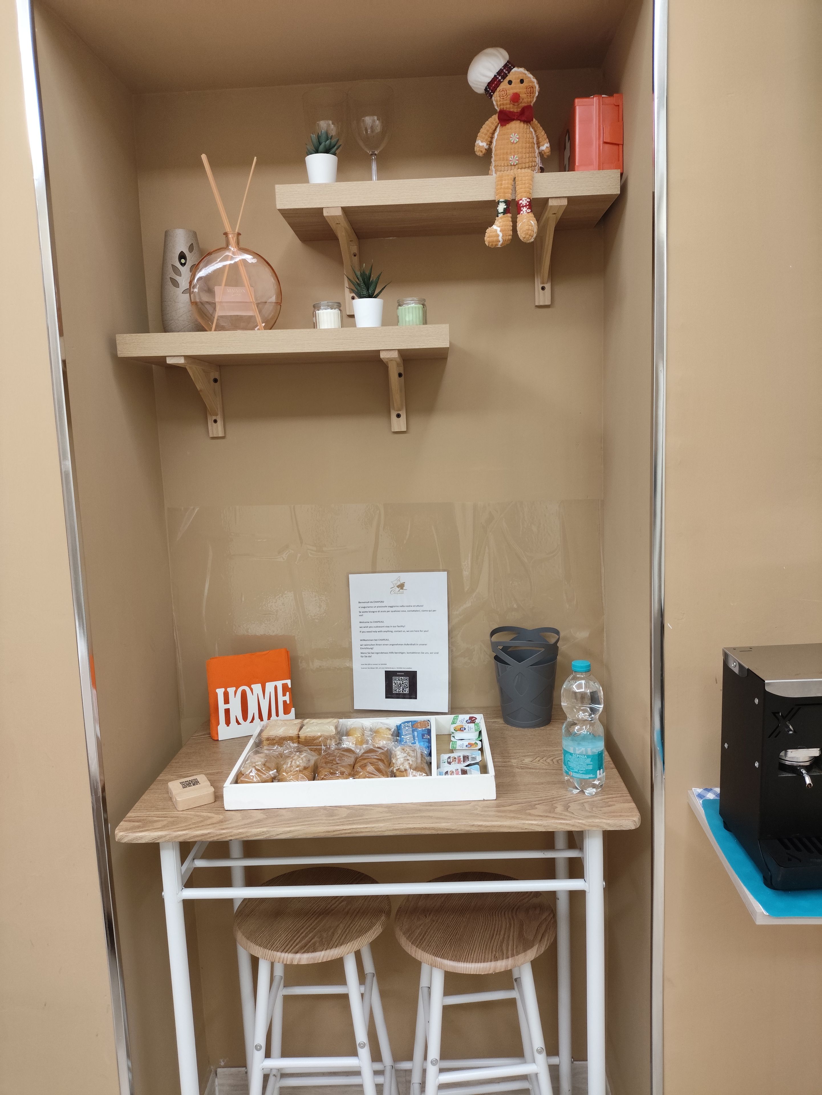 Ein kleines Zimmer mit einem Holztisch, zwei Hocker, einem kleinen Regal mit einem Teddybären, einer Topfpflanze und einem Schild mit der Aufschrift 'HOME'.