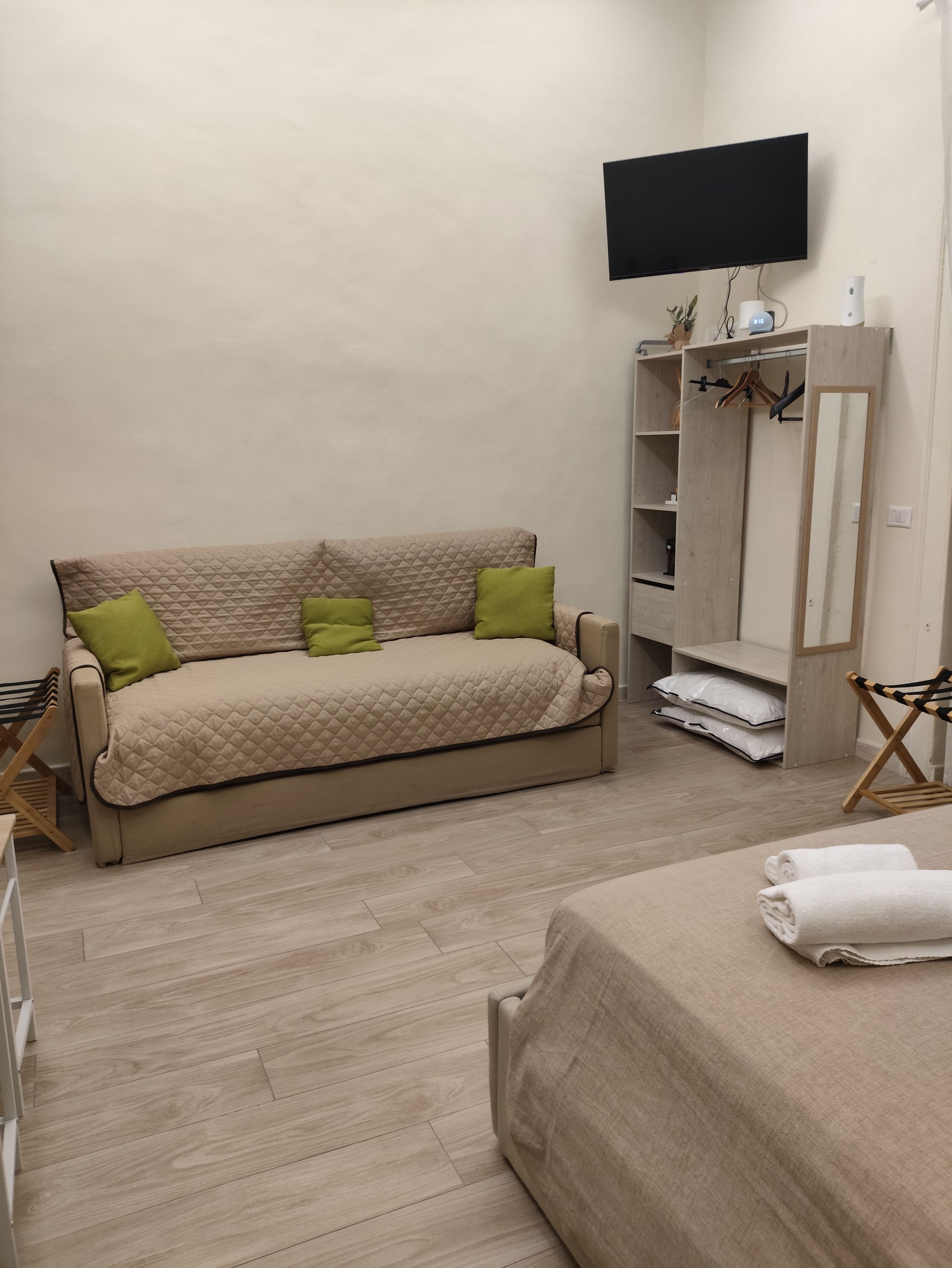 A room with a beige sofa, a television mounted on the wall, and a wooden wardrobe with shelves.