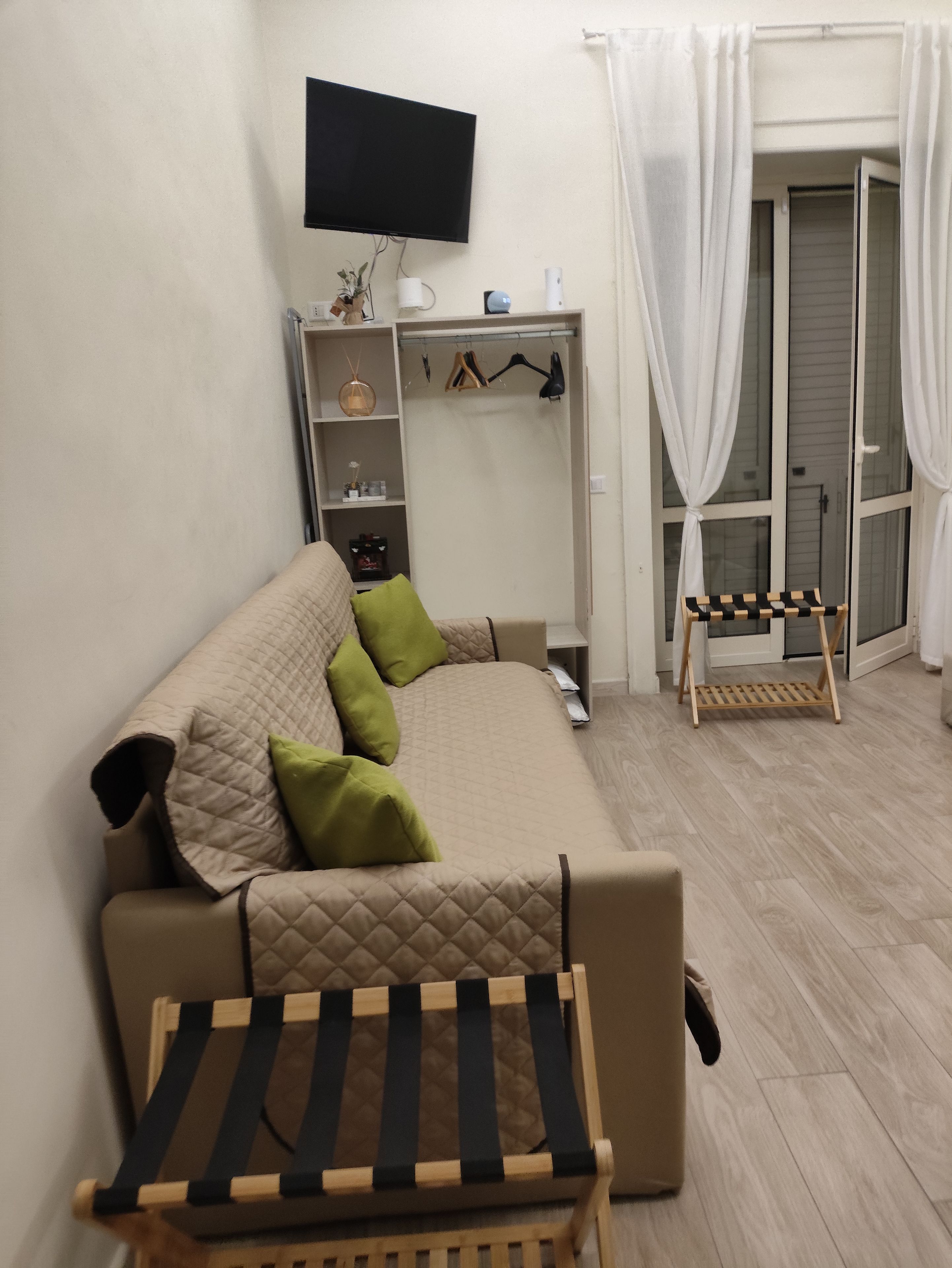 A compact living space with a sofa, a small table, a television mounted on the wall, a wardrobe, and a window with curtains.