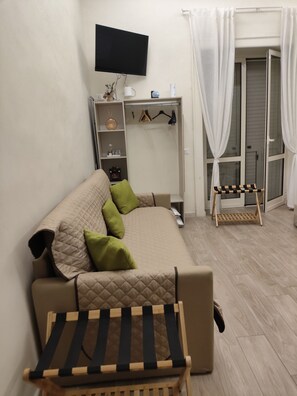 A compact living space with a sofa, a small table, a television mounted on the wall, a wardrobe, and a window with curtains.