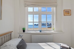 A bedroom with a bed, a window with a view of the beach, a radiator, and a small plant on the windowsill. - 1 Sunny Cove - Direct Beach Access, Sea Views (Tenby)
