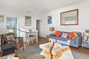 A well-lit living room with a dining area, a sofa with colorful cushions, and a large mirror on the wall. - 1 Sunny Cove - Direct Beach Access, Sea Views (Tenby)