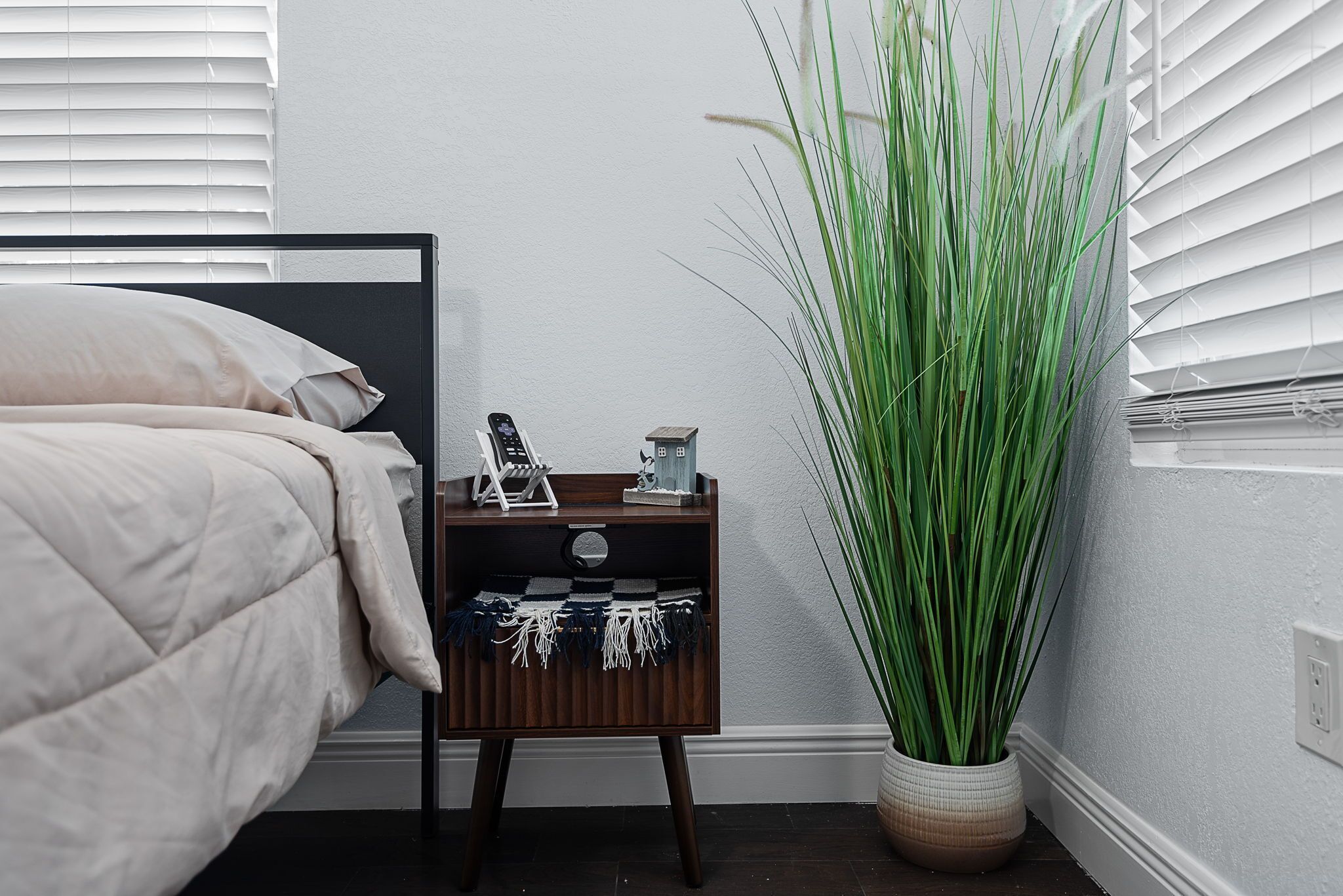 Un dormitorio con una cama, una mesita de noche con un teléfono y objetos decorativos, y una planta verde alta.