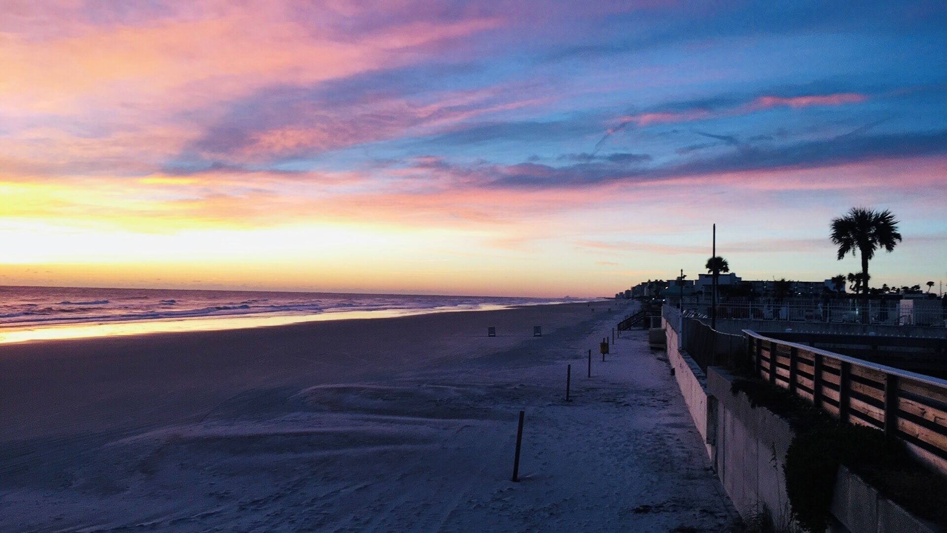 A beach with a sunset, a promenade, and palm trees.