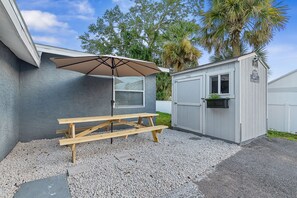 A patio area with a wooden table and a covered umbrella, a small shed, and a gravel driveway.