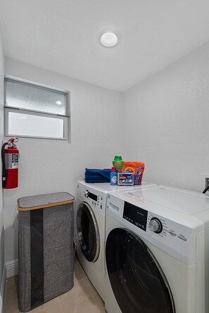 A laundry area with two washing machines, a fire extinguisher, a trash bin, and a window.