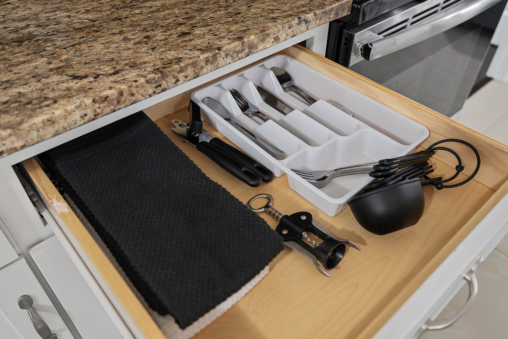 A kitchen drawer with utensils, a black cloth, and a black kitchen tool.