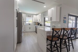 A modern kitchen with white cabinets, a marble countertop, and a bar area with stools. - Beach Escape, Private Pool ,Sleeps 8 (Fort Myers Beach)
