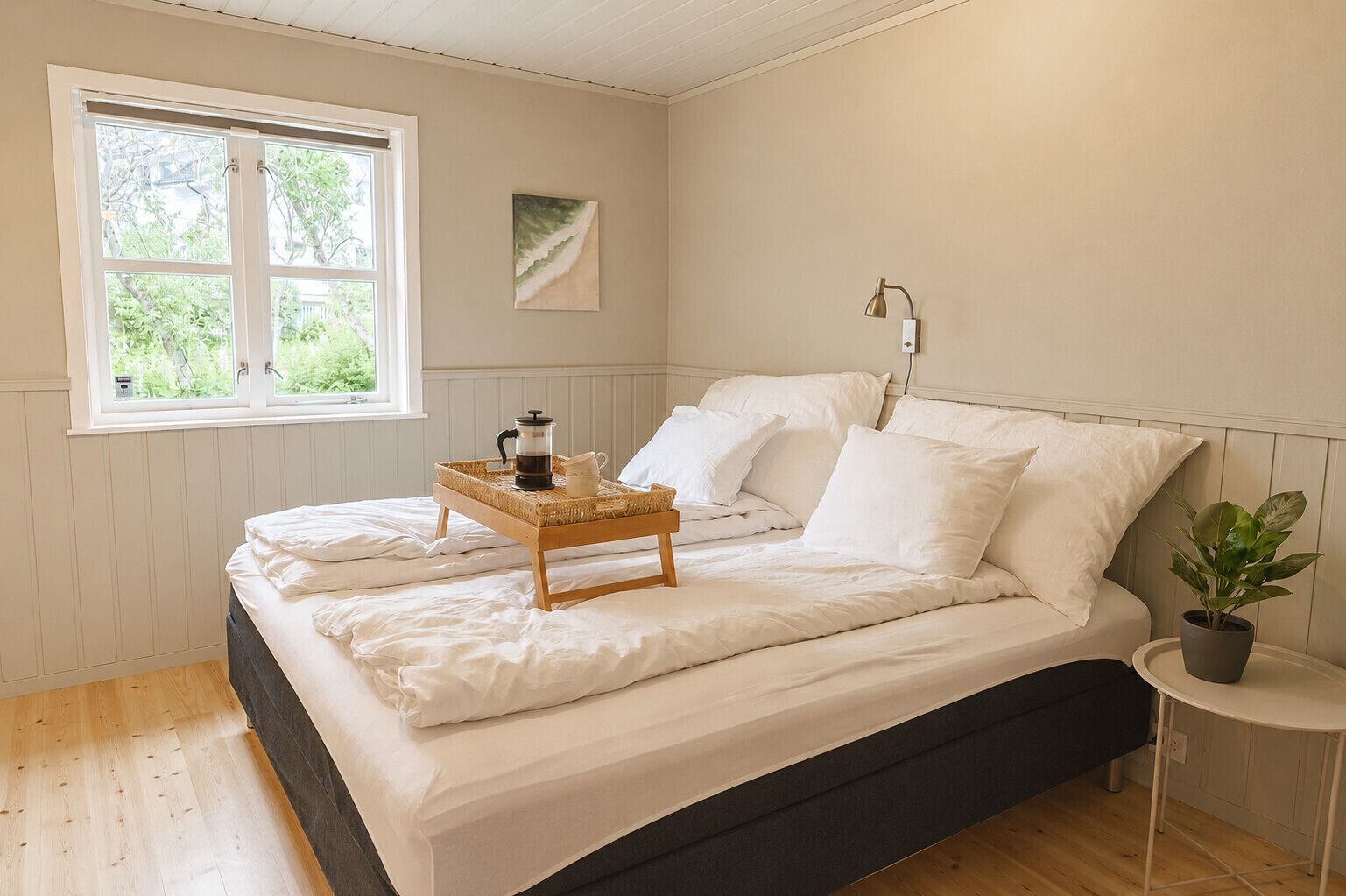 Quarto com cama, mesa de cabeceira com luminária, janela com vista para o verde e um quadro na parede.