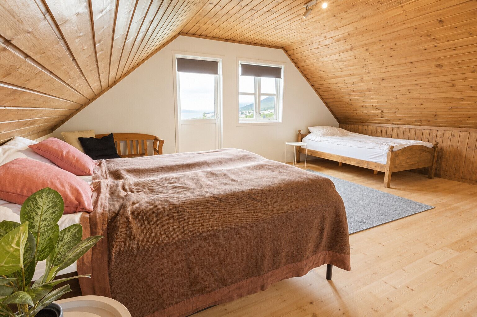 Une chambre mansardée chaleureuse, avec un plafond en pente, un grand lit recouvert d’une couverture marron, un plus petit lit, deux fenêtres et une plante en pot.