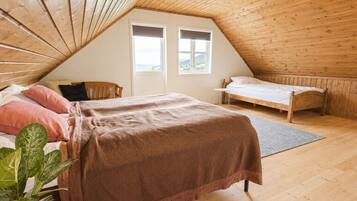 Une chambre mansardée chaleureuse, avec un plafond en pente, un grand lit recouvert d’une couverture marron, un plus petit lit, deux fenêtres et une plante en pot.