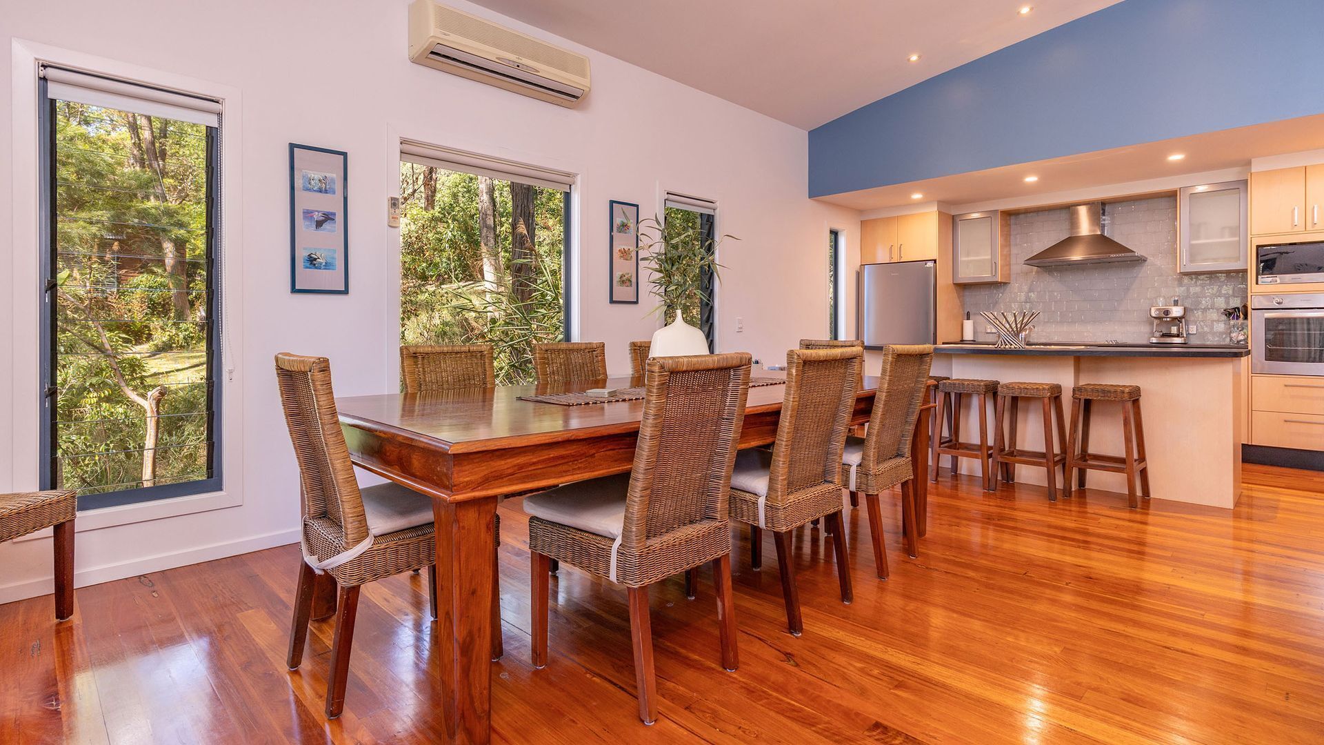 A dining area with a wooden table and wicker chairs, a kitchen with modern appliances, and large windows with a view of greenery.