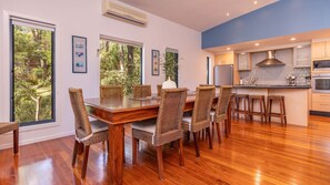Uma sala de jantar com mesa de madeira e cadeiras de vime, uma cozinha com eletrodomésticos modernos e amplas janelas com vista para o verde.