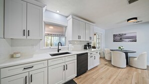 A modern kitchen with white cabinetry, a black dishwasher, and a dining area with a round table and two chairs. - Cozy Cottage Mins to Siesta Key & Downtown, Hot Tub (Sarasota)