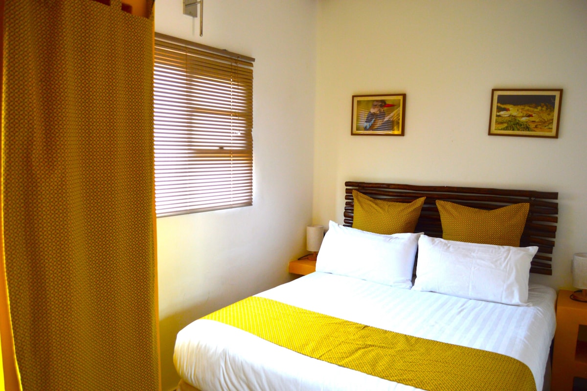 A hotel room with a bed, a window with blinds, a nightstand, and two wall-mounted paintings.
