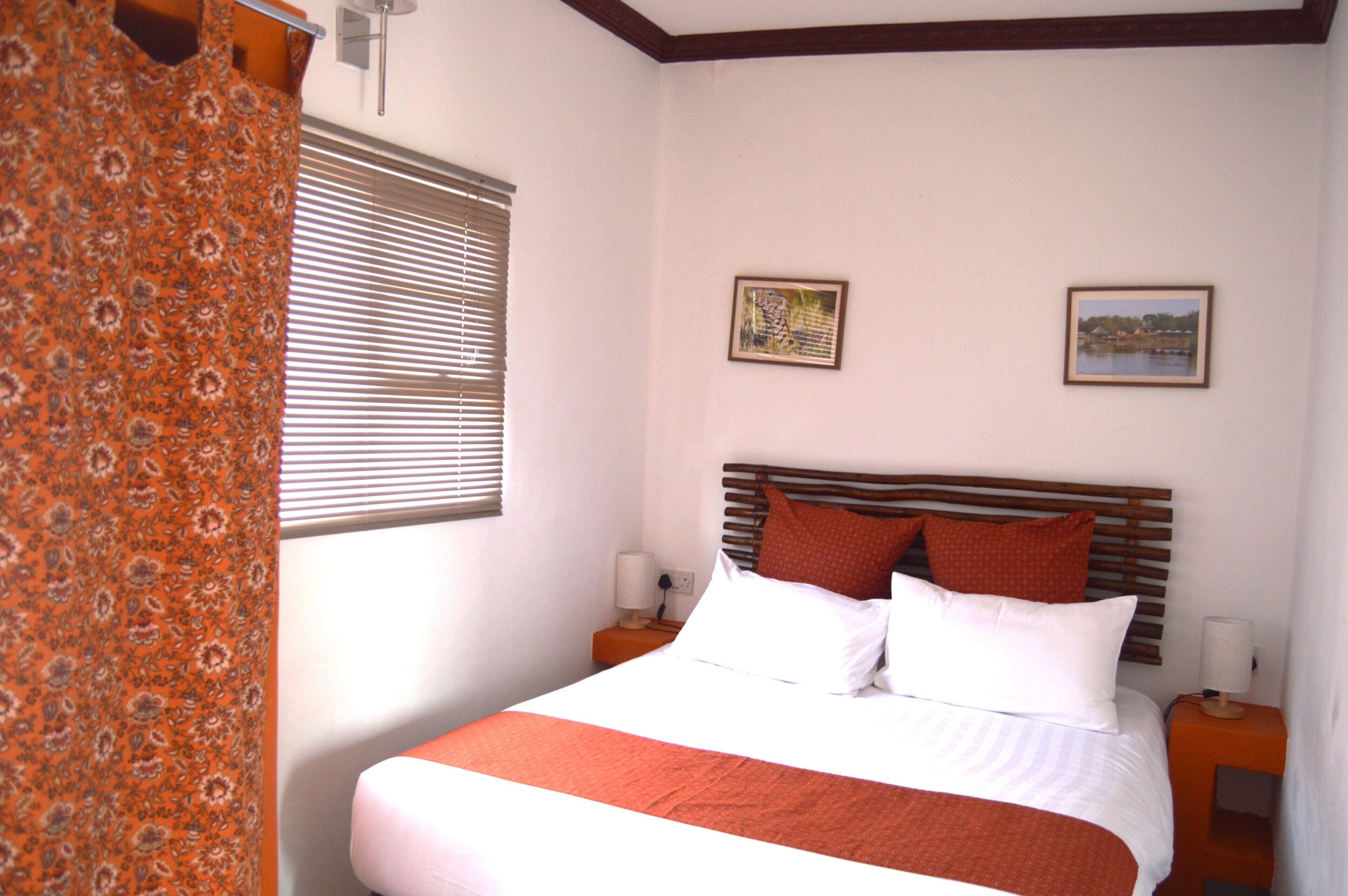 A bedroom with a bed, two bedside tables, a window with blinds, and two framed pictures on the wall.
