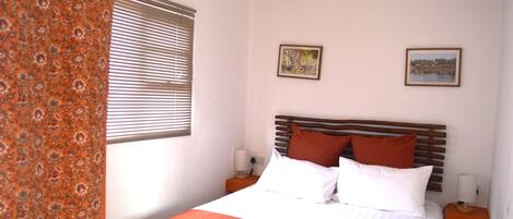 A bedroom with a bed, two bedside tables, a window with blinds, and two framed pictures on the wall.