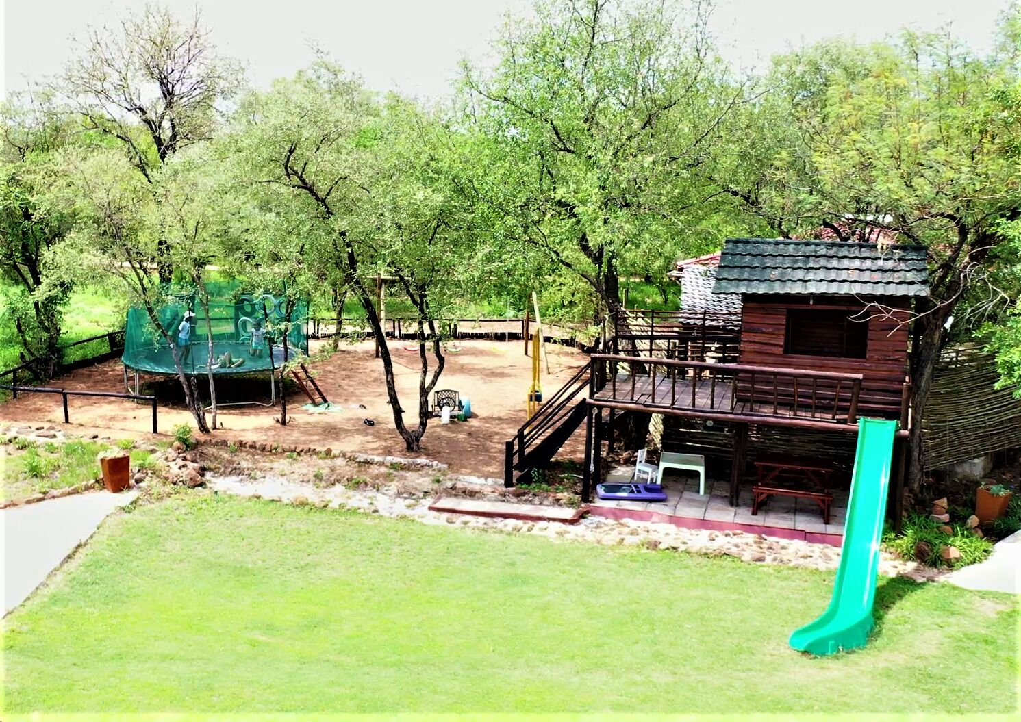 Un parque infantil con un tobogán y estructuras de escalada, rodeado de árboles y un edificio de madera con balcón.