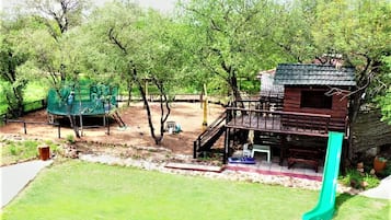 Un parque infantil con un tobogán y estructuras de escalada, rodeado de árboles y un edificio de madera con balcón.