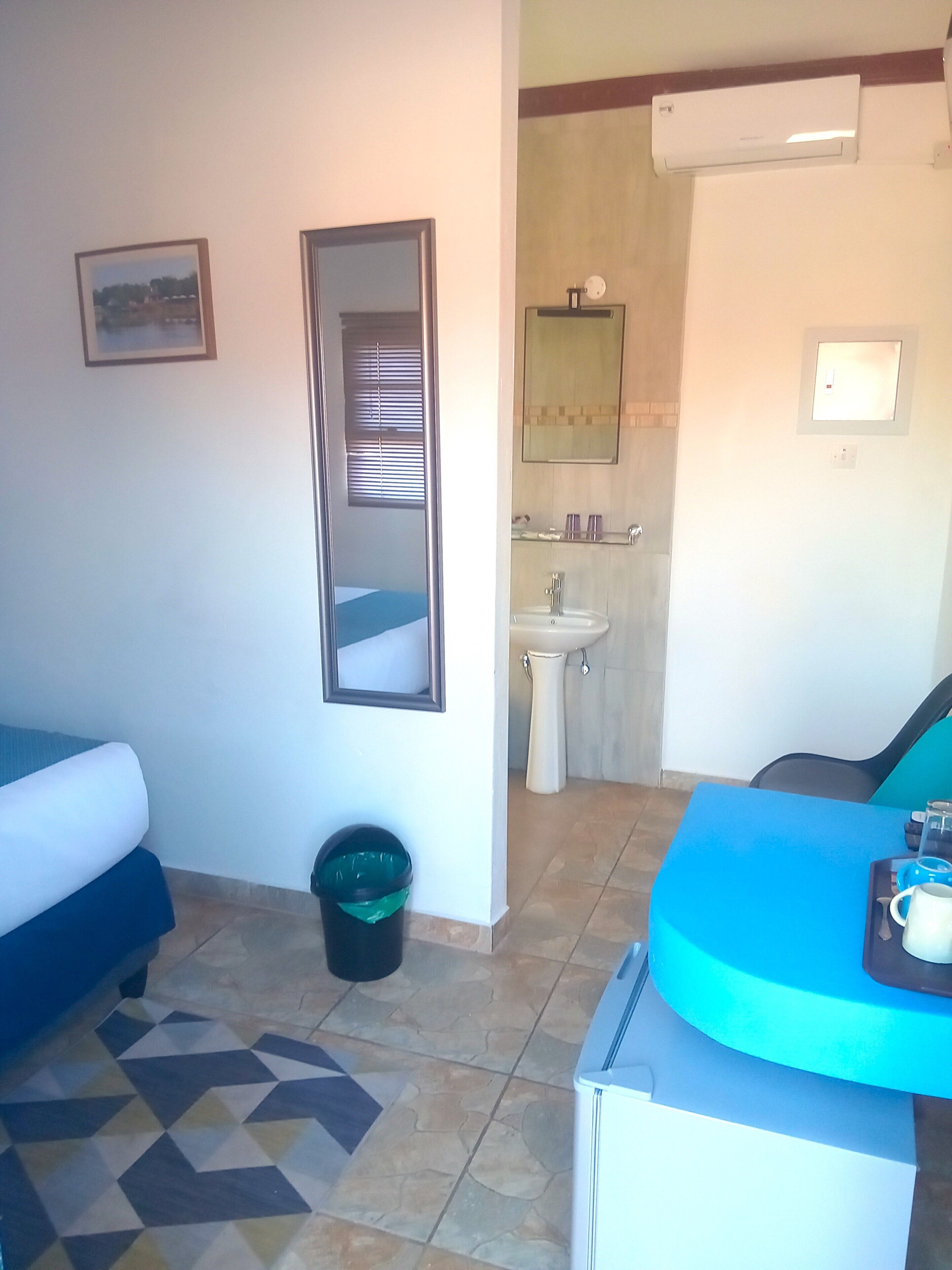 A hotel room with a bed, a desk, a chair, a trash bin, a sink, and a mirror.