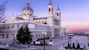 Un edificio grande e histórico con una cúpula destacada y dos torres gemelas, rodeado por una amplia plaza con faroles y algunos árboles.