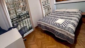 Un dormitorio con una cama, una ventana con cortinas, vista a edificios y un cuadro en la pared.