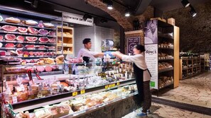 A deli counter with various meats and cheeses on display, a customer interacting with a staff member, and a sign indicating a pizza cafe.