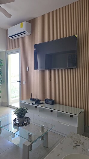 A modern living room with a flat-screen TV mounted on a wooden slat wall, a glass coffee table, and a potted plant. - Relaxing condo walking distance to Beach and all your favorites things to do  . (Boca Chica)