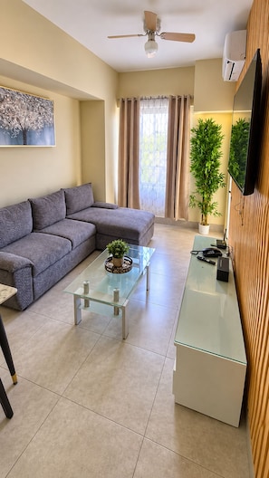 A living room with a sofa, a glass coffee table, a ceiling fan, a potted plant, and a TV mounted on the wall. - Relaxing condo walking distance to Beach and all your favorites things to do  . (Boca Chica)