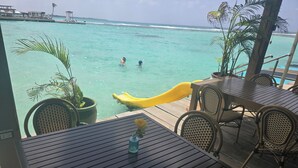 Eine Holzterrasse mit Tischen und Stühlen mit Blick auf türkisfarbenes Wasser, im Hintergrund ein geläufiger Rutsche und Palmen.