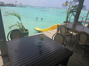 A wooden deck with tables and chairs overlooking turquoise water, with a yellow slide and palm trees in the background. - Relaxing condo walking distance to Beach and all your favorites things to do  . (Boca Chica)