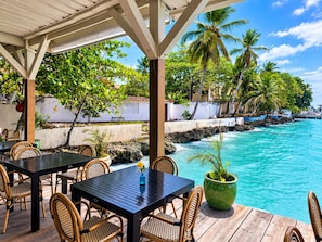 An outdoor seating area with tables and chairs, overlooking a turquoise ocean and palm trees. - Relaxing condo walking distance to Beach and all your favorites things to do  . (Boca Chica)