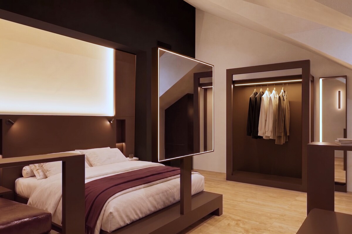 A modern hotel room with a bed, a mirror, a closet with clothes, and a small table.