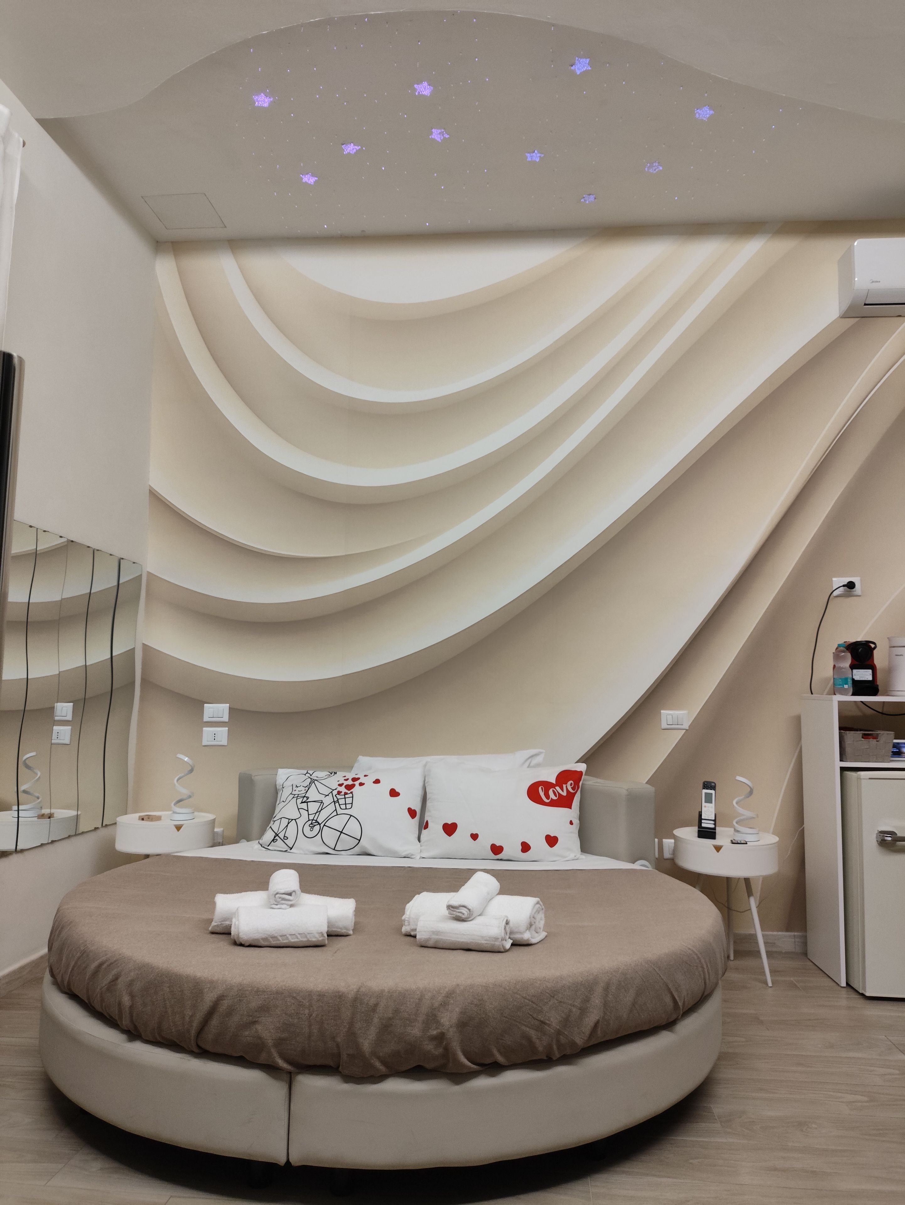 A modern bedroom with a curved bed, a ceiling projector, and a bedside table with a remote control.