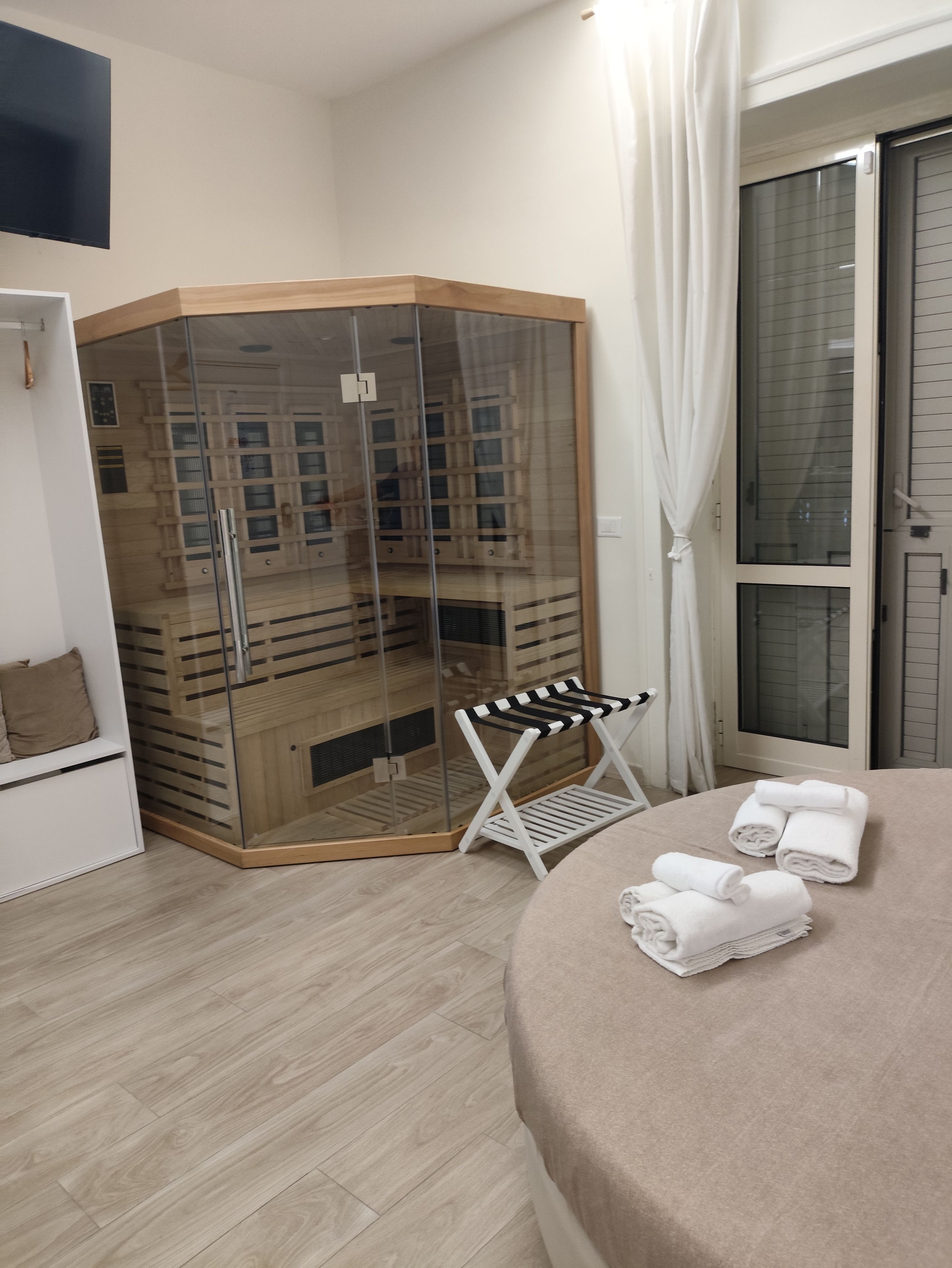 Camera d'albergo con cabina doccia in vetro, una panca bianca e asciugamani ordinatamente piegati sul letto.