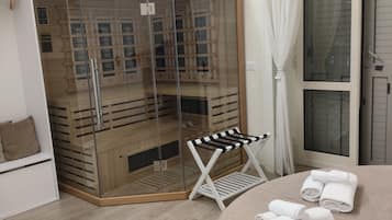 Camera d'albergo con cabina doccia in vetro, una panca bianca e asciugamani ordinatamente piegati sul letto.