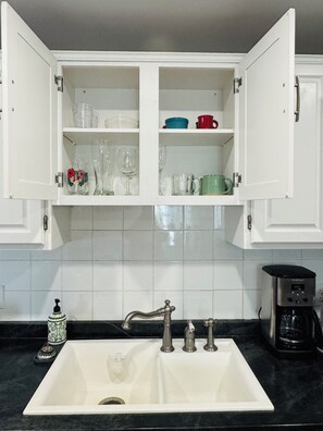 Eine Küche mit einem weißen Spülbecken, zwei Wasserhähnen, einer Kaffeemaschine und einem Schrank mit Gläsern und Geschirr.