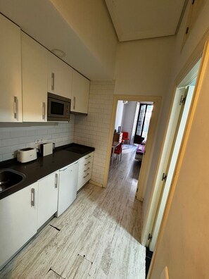A kitchen with white cabinets, a microwave, and a toaster. There is a wooden floor and a doorway leading to a room with a bed and a chair. - PUERTA DEL SOL CARRETAS APARTMENTS&ROOMS (Madrid)